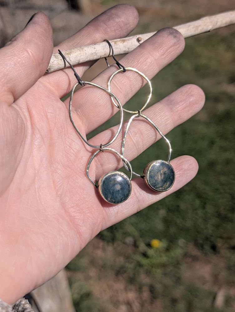 Asymmetric linked metal loops with round green patterned stones, displayed in a hand.