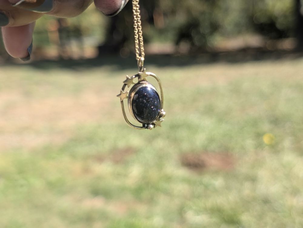 Sparkling dark blue oval pendant surrounded by an open metal frame with small stars and rounded details.