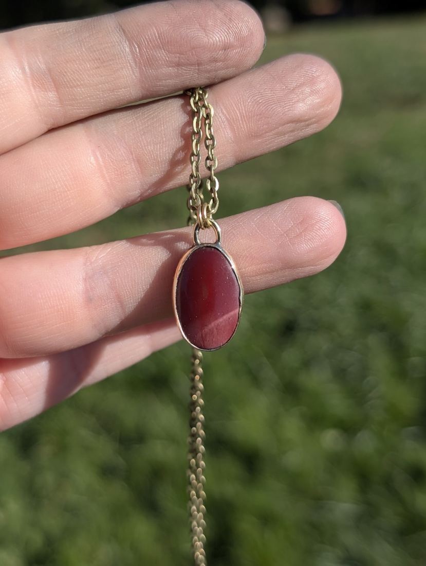 Oval red jasper pendant held in a hand outdoors.