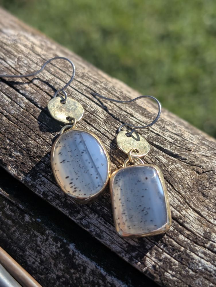 An oval and a rectangular speckled stone below round brass links with niobium ear hooks, resting on wood.