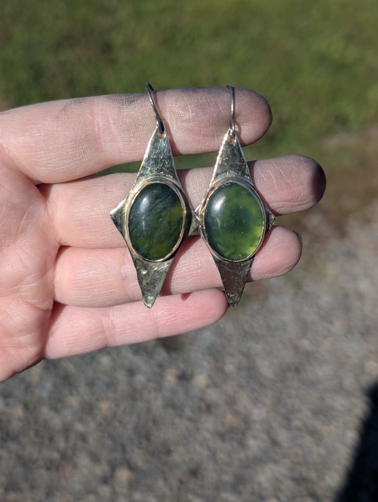 Oval green nephrite jade stones in elongated pointed metal settings with niobium ear hooks, held in a hand.