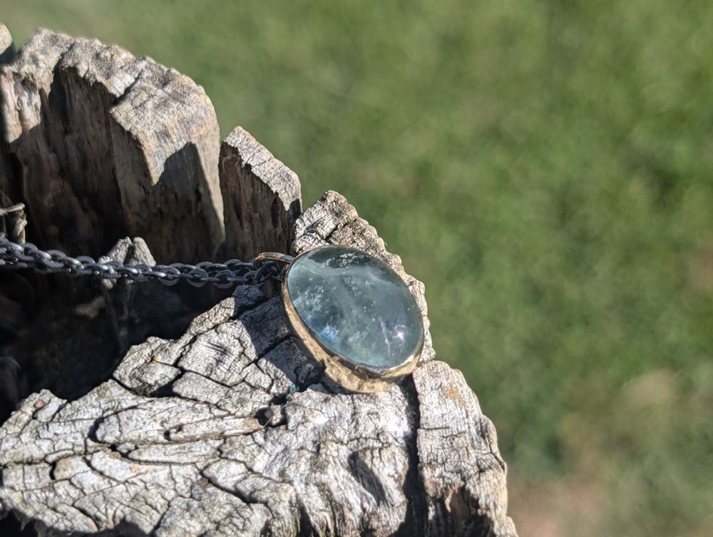 Pale green oval stone pendant in a handmade metal setting, resting on weathered wood.