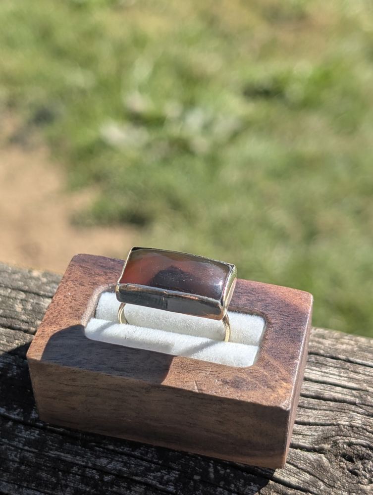 Rectangular red-and-dark patterned stone ring displayed in a wooden ring box.